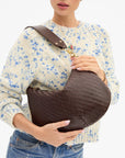 A woman wearing a cream knit sweater with blue floral patterns and blue jeans holds a dark brown woven shoulder bag with a wide brown strap and gold-tone hardware against a plain white background.