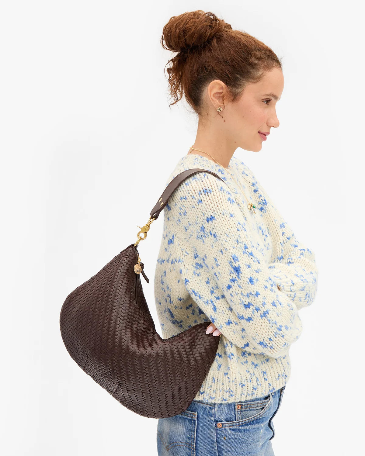 A woman in a cream knit sweater with blue speckles and jeans is shown from the side, carrying a large brown woven crescent shoulder bag with gold-tone hardware against a plain white background.
