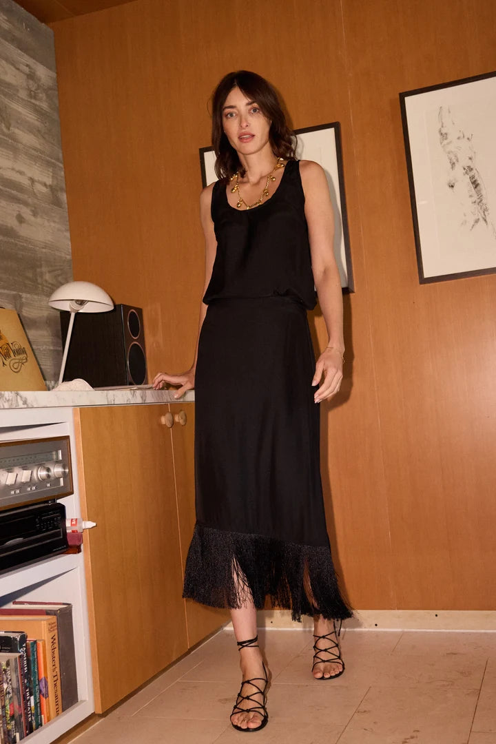 A woman models a black sleeveless midi skirt with a fringe hem, paired with a black top and strappy sandals, standing in a room with warm wood-paneled walls.
