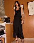 A woman models a black sleeveless midi skirt with a fringe hem, paired with a black top and strappy sandals, standing in a room with warm wood-paneled walls.