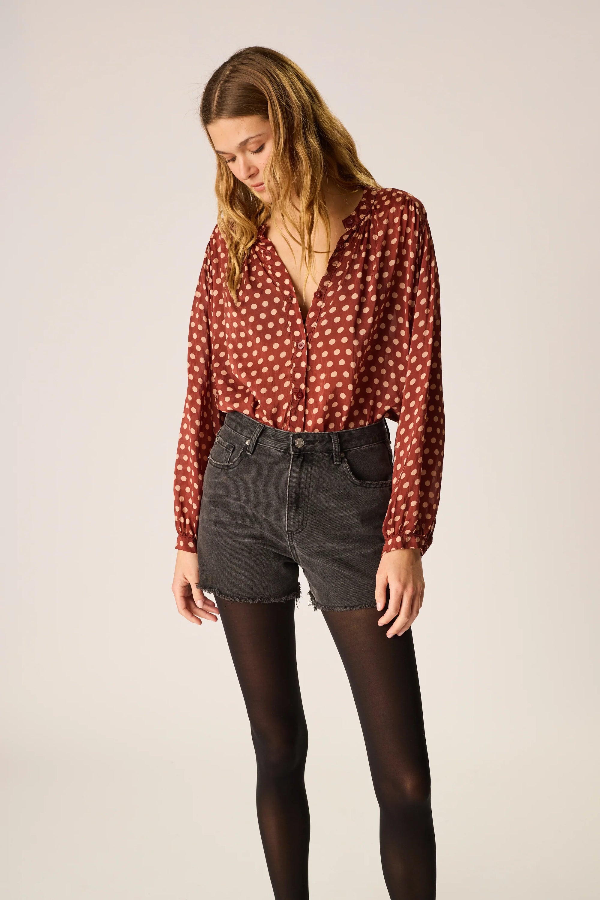 Woman wearing a red polka dot blouse and black jeans on a plain background