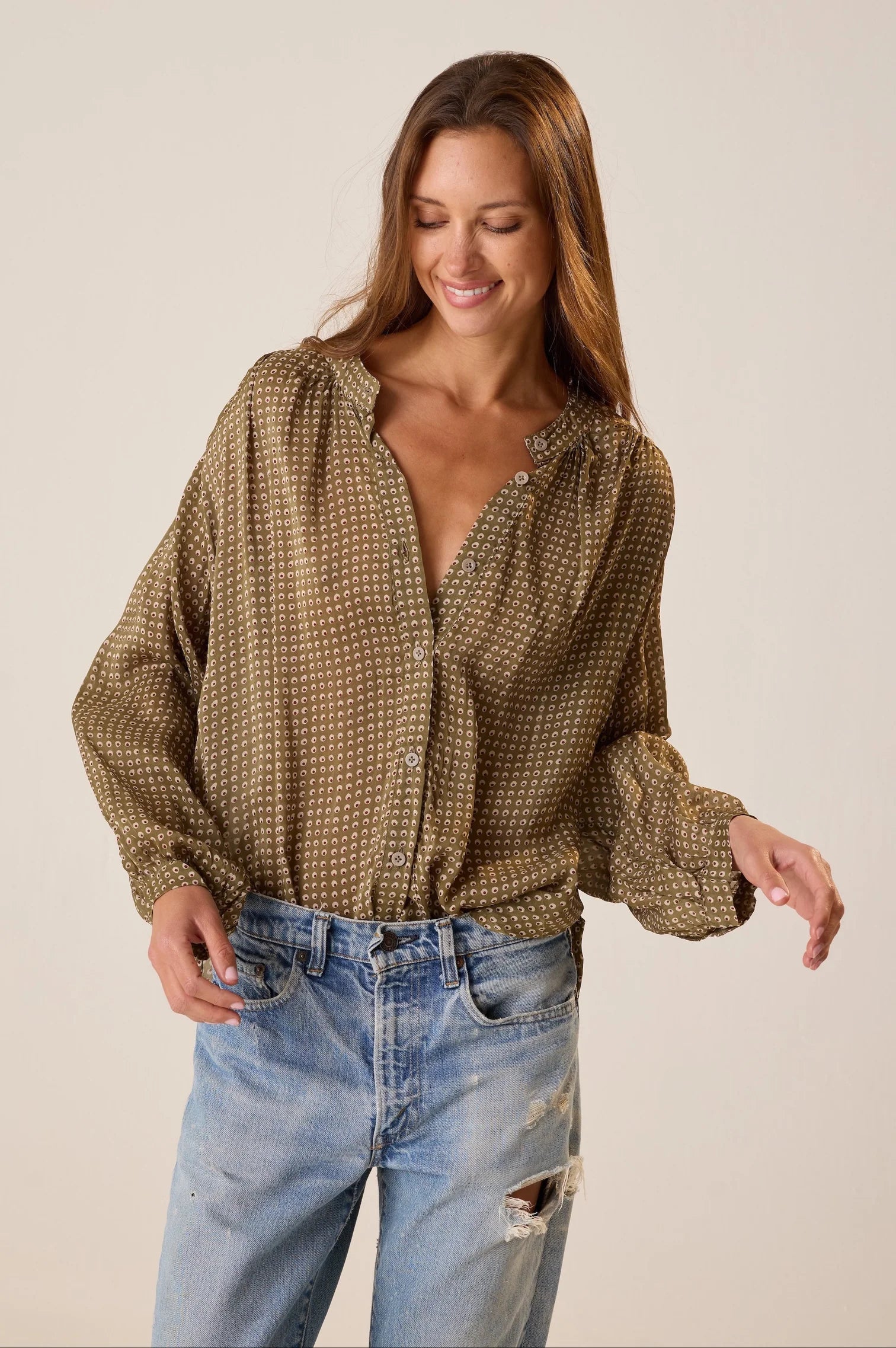 A woman wearing a brown and beige patterned long-sleeve blouse with a split neckline and button front, paired with light-wash distressed jeans, against a plain beige studio background.