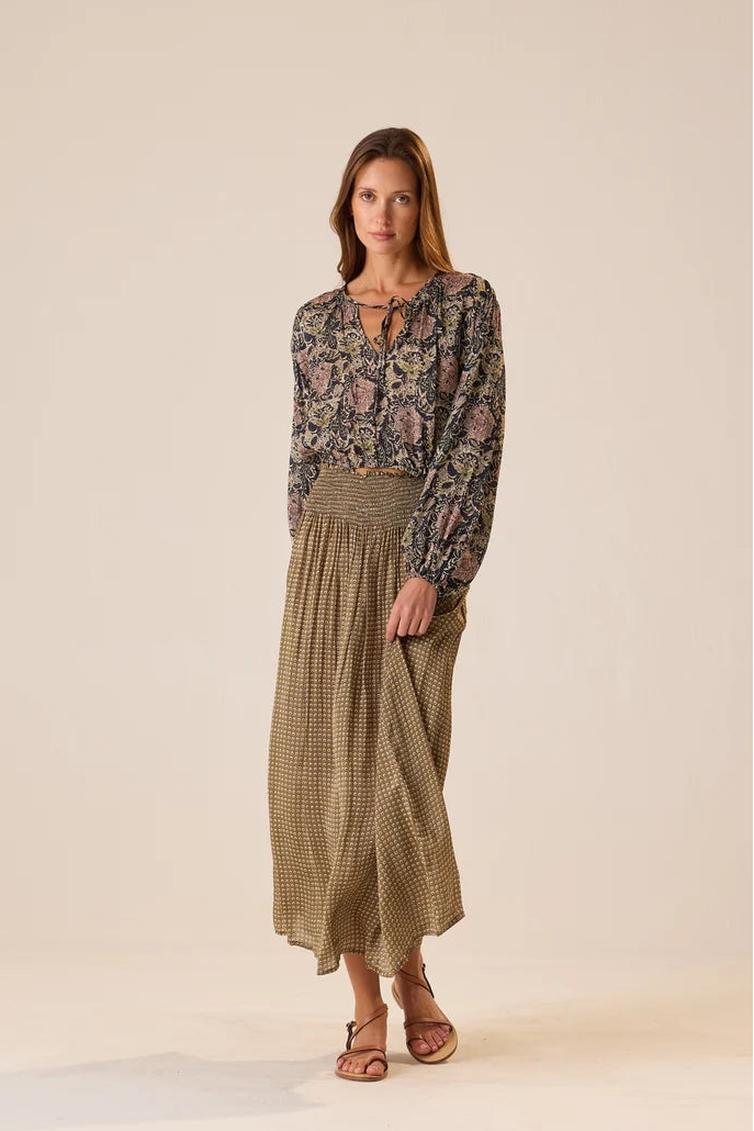A woman modeling a long-sleeve bohemian floral print blouse paired with a high-waisted, pleated olive green and beige gingham maxi skirt, set against a solid warm beige studio background.