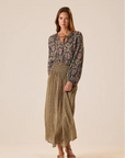 A woman modeling a long-sleeve bohemian floral print blouse paired with a high-waisted, pleated olive green and beige gingham maxi skirt, set against a solid warm beige studio background.
