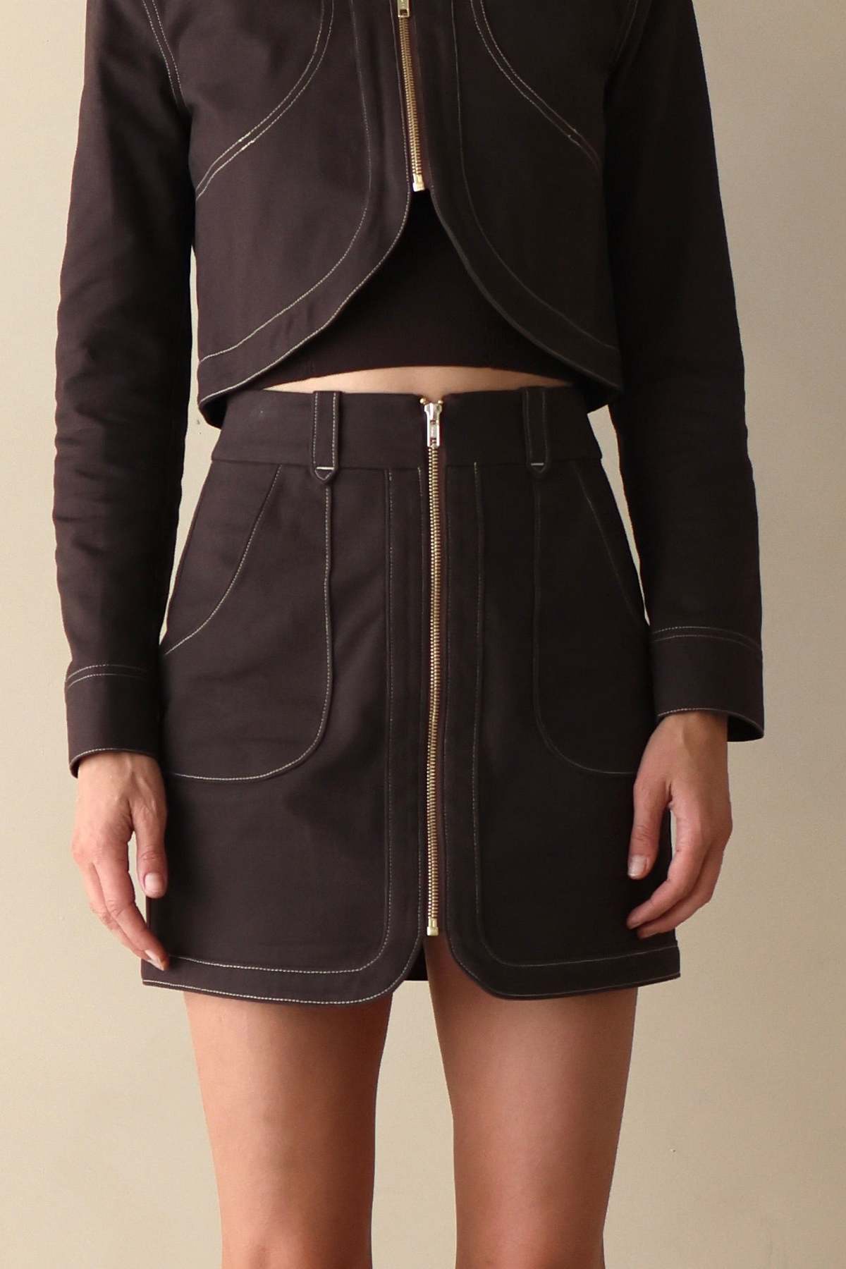 A model wearing a dark brown cropped jacket and matching high-waisted mini skirt with contrast light stitching and a front zipper, set against a plain beige background.