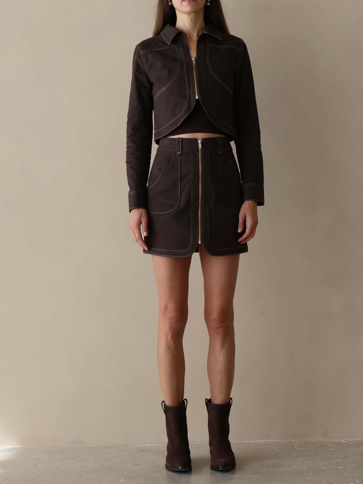 A model stands against a plain beige studio backdrop wearing a dark brown cropped zip-front jacket and matching mini skirt set, paired with dark ankle boots.