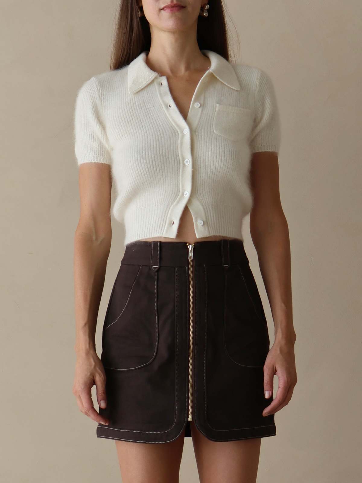 Woman wearing a white cropped knit shirt and brown shorts against a beige background