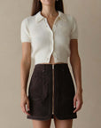 Woman wearing a white cropped knit shirt and brown shorts against a beige background