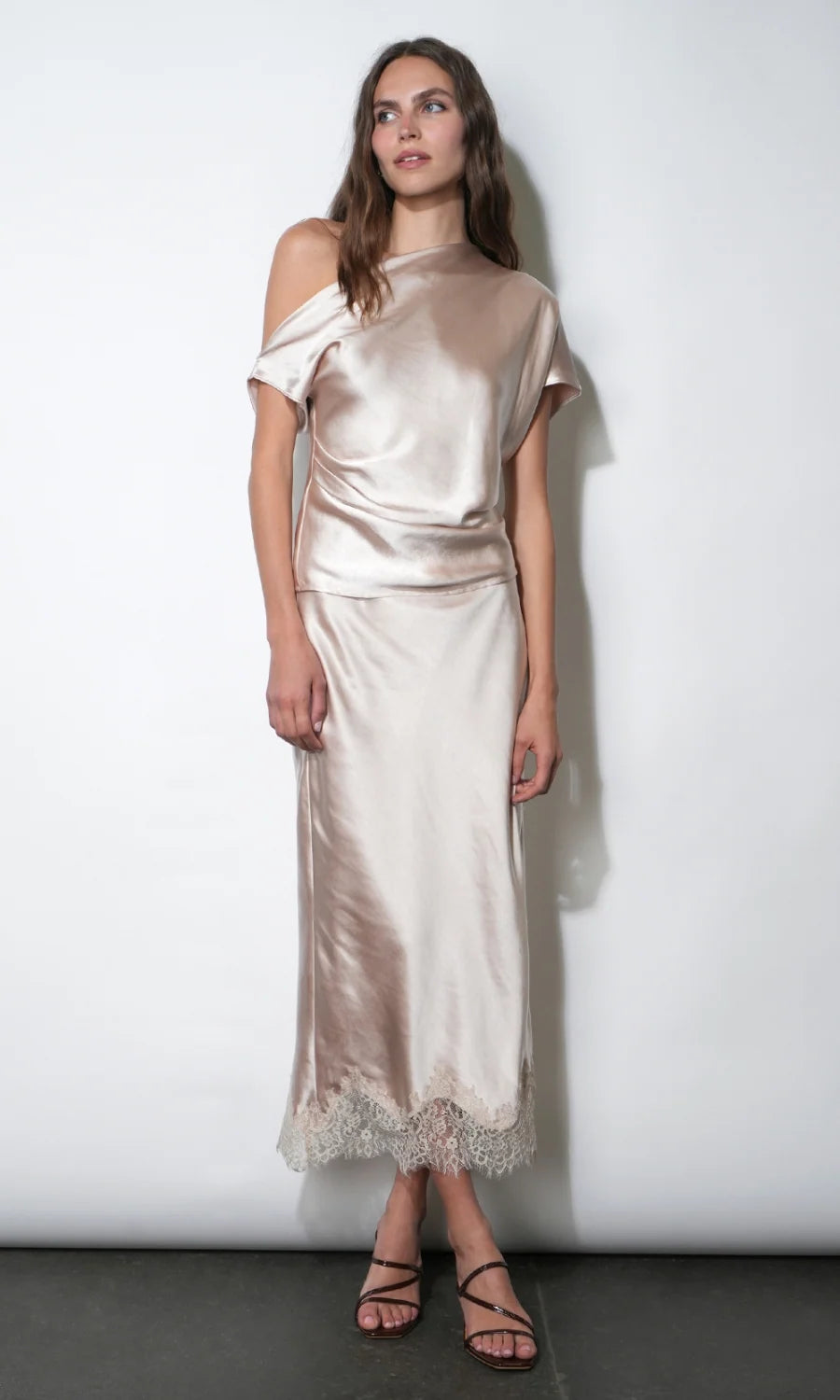A woman modeling a champagne satin two-piece set consisting of a cold-shoulder top and a matching midi skirt with a lace hem, set against a plain white studio background.
