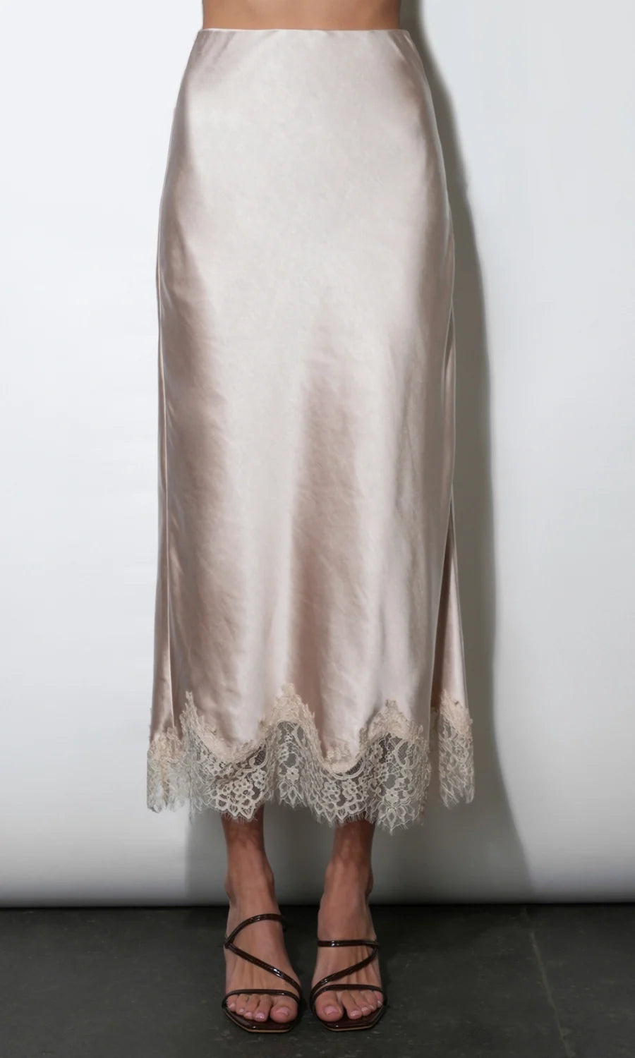 A light beige satin midi skirt with a scalloped lace hem, shown on a model wearing black strappy heels against a plain white wall.
