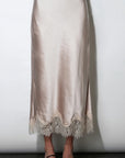 A light beige satin midi skirt with a scalloped lace hem, shown on a model wearing black strappy heels against a plain white wall.