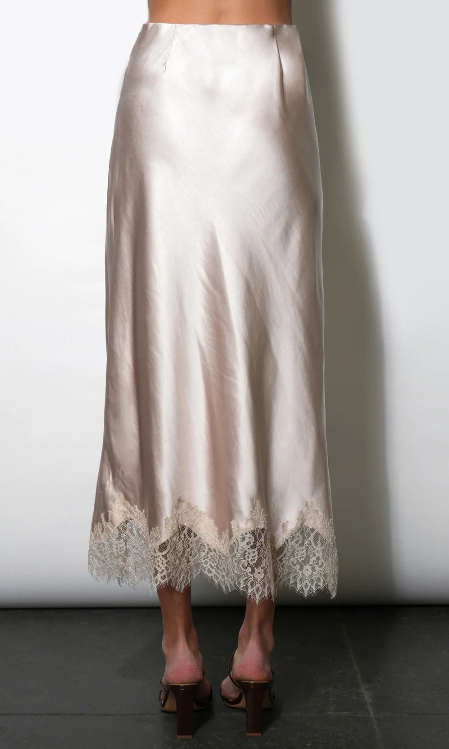 A satin midi skirt in a champagne beige color with a lace hem, shown on a model from the back against a white wall.