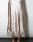 A satin midi skirt in a champagne beige color with a lace hem, shown on a model from the back against a white wall.