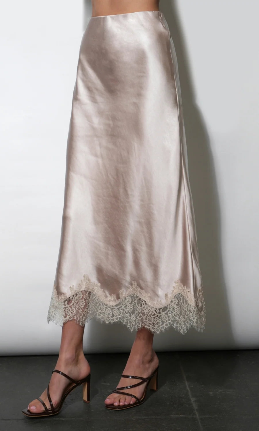 A satin midi skirt in a champagne beige color with a lace hem, shown on a model wearing black strappy mule heels against a plain light gray wall.