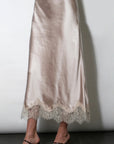 A satin midi skirt in a champagne beige color with a lace hem, shown on a model wearing black strappy mule heels against a plain light gray wall.