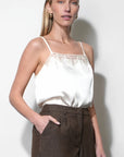Woman wearing a white satin camisole with lace trim and brown pants against a white background