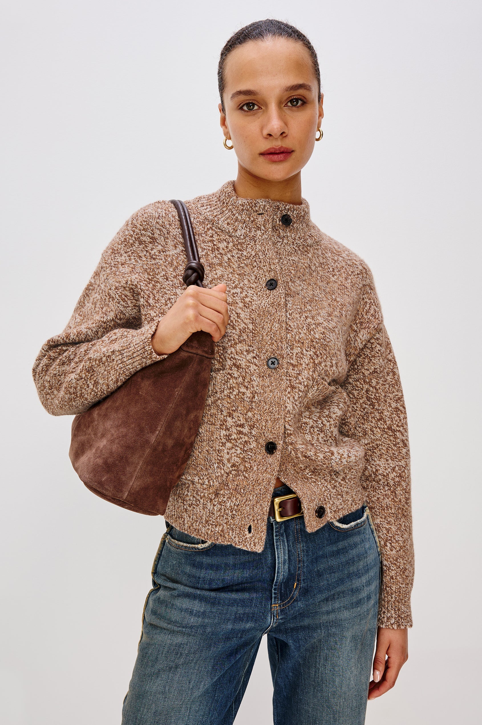 Woman wearing a brown patterned cardigan and blue jeans, holding a brown bag against a white background