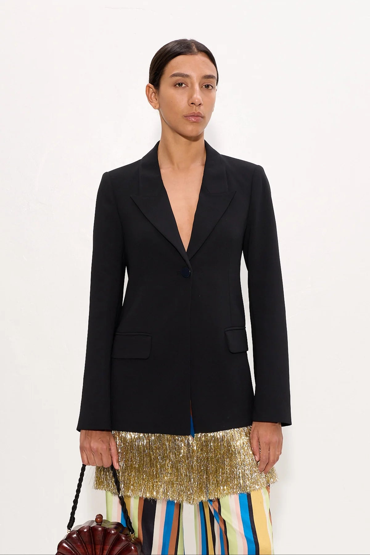 A model wearing a black structured blazer over a colorful striped skirt with a gold fringe hem, holding a small brown handbag against a plain white background.
