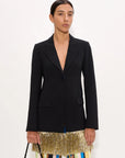A model wearing a black structured blazer over a colorful striped skirt with a gold fringe hem, holding a small brown handbag against a plain white background.