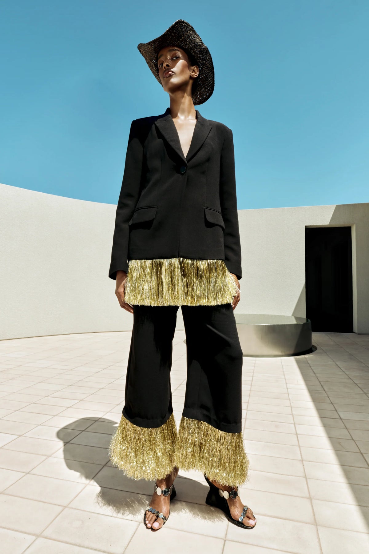 A model stands outdoors in a minimalist concrete setting under a clear blue sky, wearing a black blazer and wide-leg pants with metallic gold fringe detailing at the hem and upper leg, paired with a black hat and sandals.