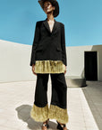 A model stands outdoors in a minimalist concrete setting under a clear blue sky, wearing a black blazer and wide-leg pants with metallic gold fringe detailing at the hem and upper leg, paired with a black hat and sandals.