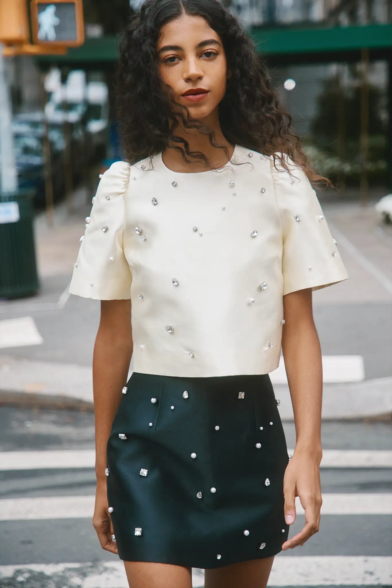 Woman wearing a white top with embellishments and a dark green skirt with white patterns on a city street.