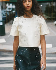 Woman wearing a white top with embellishments and a dark green skirt with white patterns on a city street.