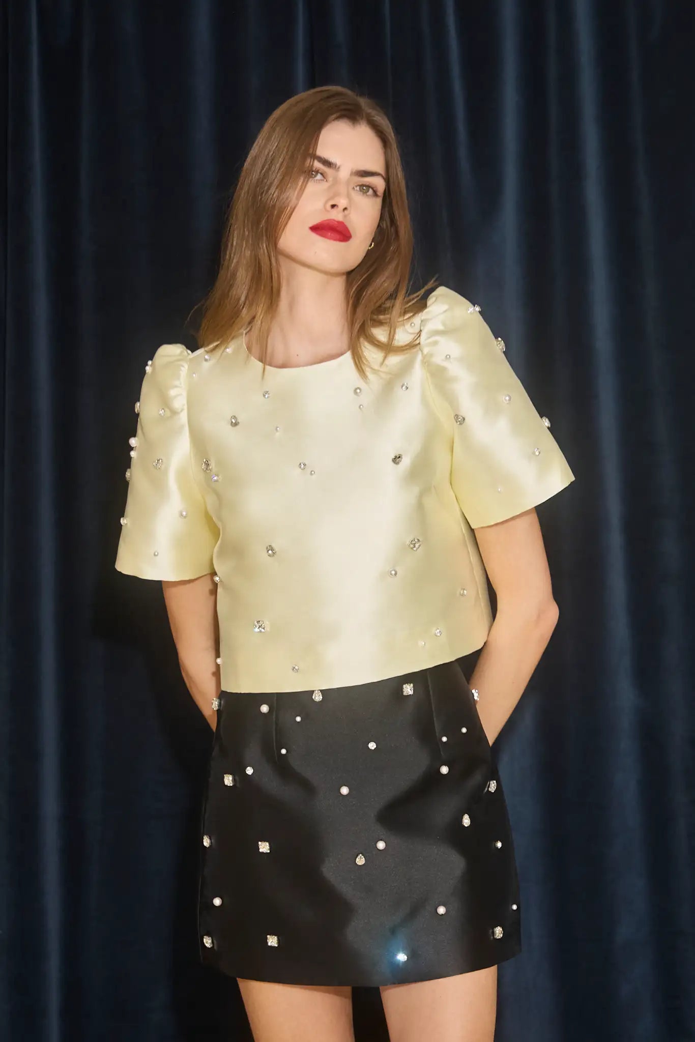 Woman wearing a light yellow top with embellishments and a black skirt with studs against a dark curtain background.
