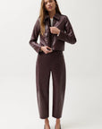 A woman modeling a dark brown faux leather jacket and matching wide-leg cropped trousers against a plain light gray studio background.