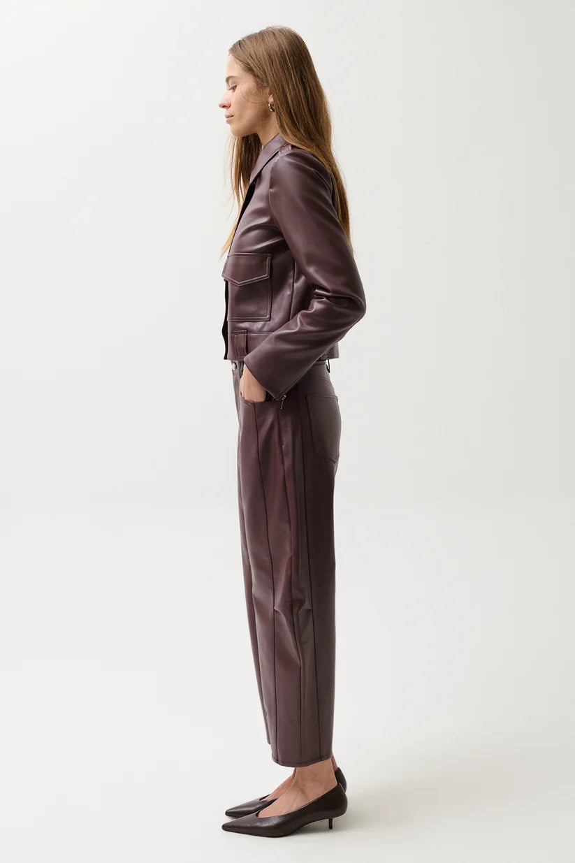 A woman stands in profile wearing a matching dark plum faux-leather blazer and wide-leg trousers set, paired with black pointed-toe flats, against a plain light gray studio background.