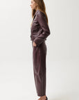 A woman stands in profile wearing a matching dark plum faux-leather blazer and wide-leg trousers set, paired with black pointed-toe flats, against a plain light gray studio background.