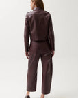 A woman viewed from the back wearing a dark plum faux-leather cropped jacket and matching wide-leg cropped pants, paired with dark pointed-toe heels, against a light gray studio background.