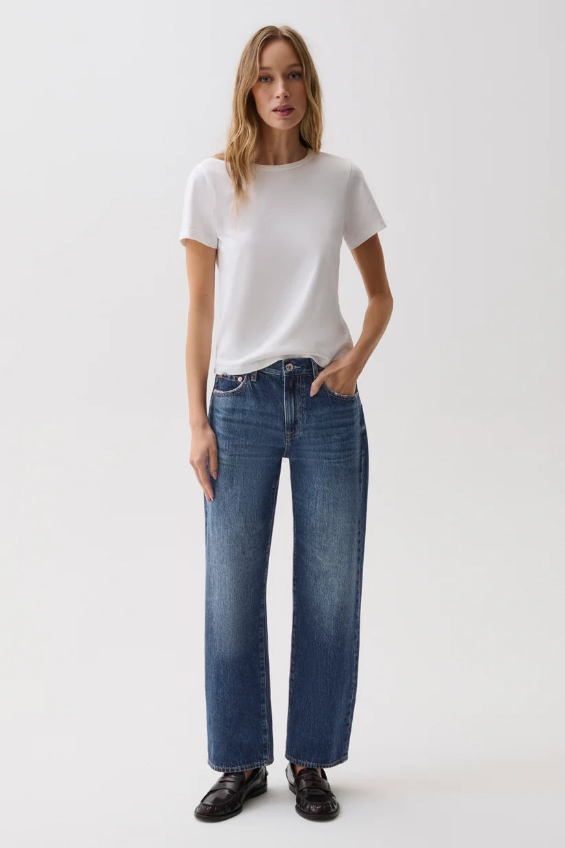A woman modeling a white short-sleeve crewneck t-shirt and high-waisted, wide-leg blue jeans with dark brown leather loafers, shot against a seamless white studio background.