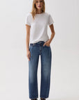 A woman modeling a white short-sleeve crewneck t-shirt and high-waisted, wide-leg blue jeans with dark brown leather loafers, shot against a seamless white studio background.