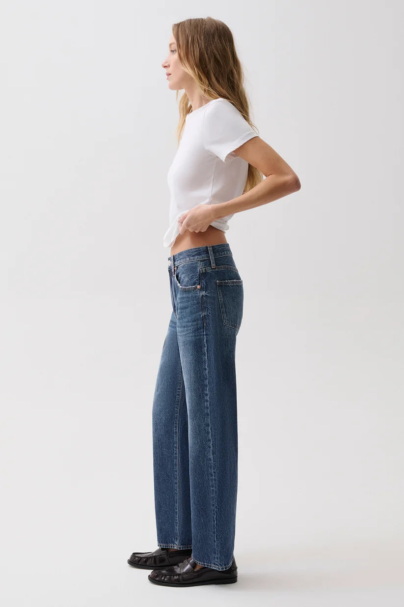 A woman in a white t-shirt and blue wide-leg jeans stands in profile against a plain light gray studio background.