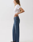 A woman in a white t-shirt and blue wide-leg jeans stands in profile against a plain light gray studio background.