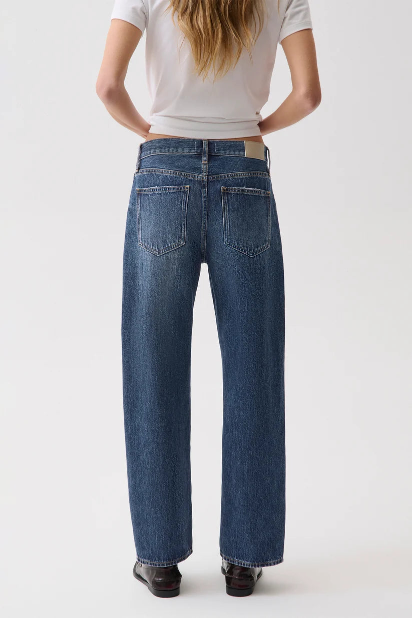A model viewed from the back wearing a pair of wide-leg blue jeans, a white t-shirt, and black shoes, set against a plain light gray studio background.