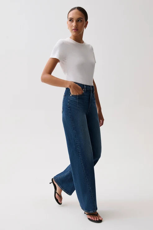 A woman modeling a white short-sleeve top and high-waisted, wide-leg blue denim jeans against a plain light gray studio background.