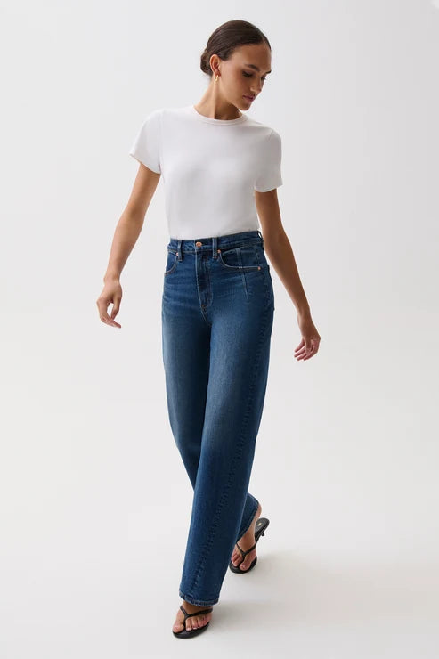 A model wearing a white short-sleeve crewneck t-shirt, high-waisted wide-leg blue jeans with a gold-tone belt, and black heeled sandals, standing against a plain light gray studio background.