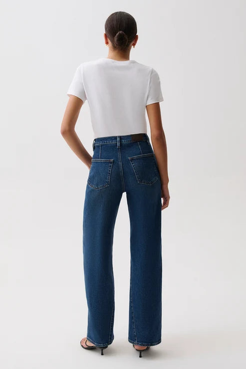 A model wearing a white short-sleeve crewneck t-shirt, high-waisted wide-leg blue jeans with a gold-tone belt, and black heeled sandals, standing against a plain light gray studio background, back