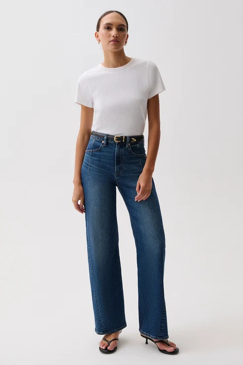 A model wearing a white short-sleeve crewneck t-shirt, high-waisted wide-leg blue jeans with a gold-tone belt, and black heeled sandals, standing against a plain light gray studio background.
