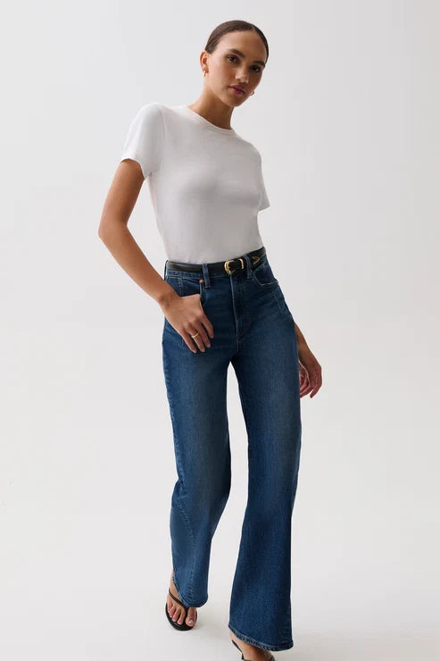 A model wearing a white short-sleeve crewneck t-shirt, high-waisted wide-leg blue jeans with a gold-tone belt, and black heeled sandals, standing against a plain light gray studio background.