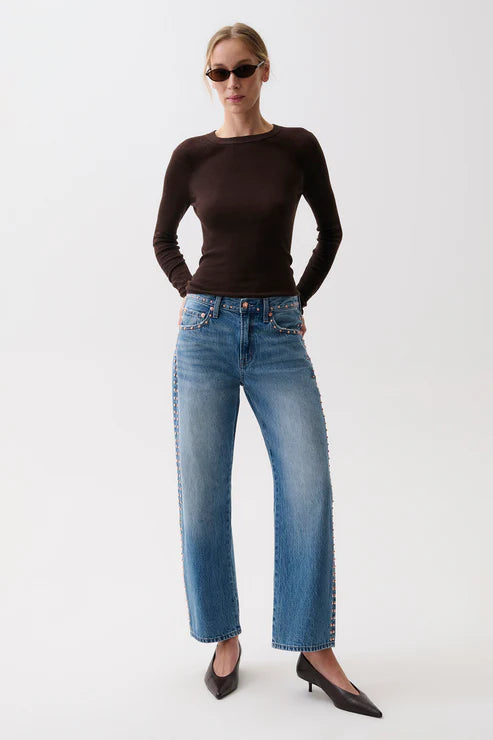 A woman modeling a dark brown long-sleeve knit top and wide-leg blue jeans with decorative silver studs down the sides, paired with black pointed-toe heels, against a plain white studio background.