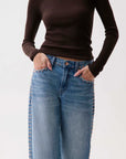A model wearing a dark brown ribbed long-sleeve top and light blue jeans with gold-tone stud detailing along the outer seams of the legs, standing against a plain white background.