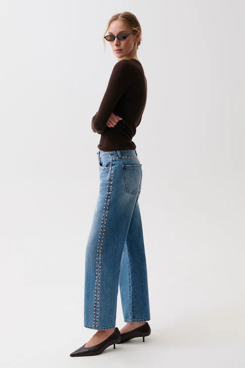 A woman wearing blue wide-leg jeans with silver chain detailing down the side, a dark brown long-sleeve top, and black pointed-toe heels, standing against a plain white studio background.