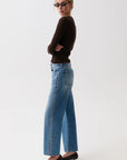 A woman wearing blue wide-leg jeans with silver chain detailing down the side, a dark brown long-sleeve top, and black pointed-toe heels, standing against a plain white studio background.
