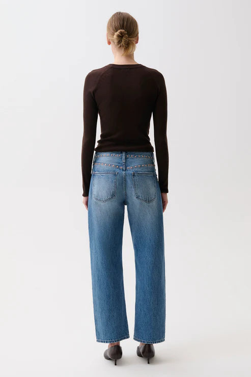 A woman viewed from behind wearing a dark brown long-sleeve top, blue wide-leg jeans, and black heels against a plain light gray studio background.