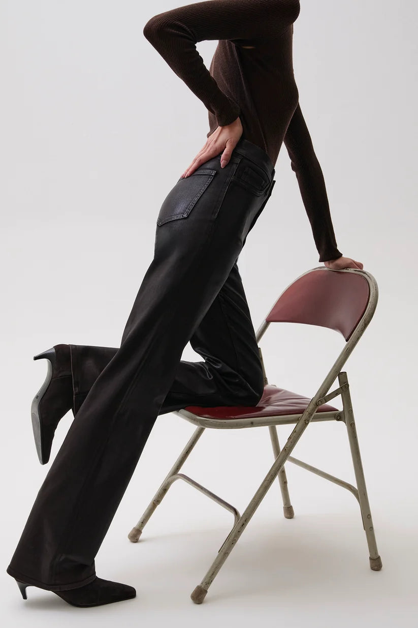 A model wearing black leather flare pants, a dark brown long-sleeve top, and black pointed-toe heels poses next to a vintage folding chair with a burgundy seat and back, set against a plain white studio background.