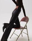 A model wearing black leather flare pants, a dark brown long-sleeve top, and black pointed-toe heels poses next to a vintage folding chair with a burgundy seat and back, set against a plain white studio background.