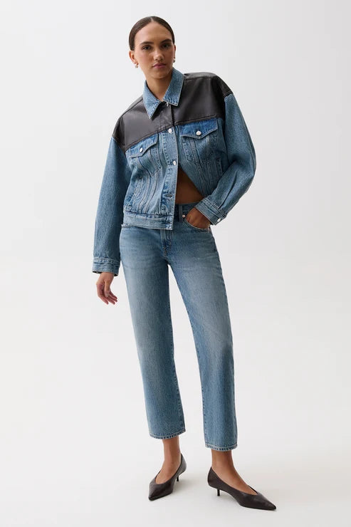 A model wearing a cropped denim jacket with a color-blocked design in medium and dark blue, paired with matching blue jeans and black pointed-toe heels, against a plain white studio background.